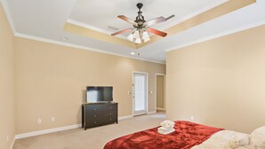 A bedroom with beige walls, a ceiling fan, a dark dresser, a bed with a red blanket, and a door leading to another room. - Large spacious Home (Enterprise)