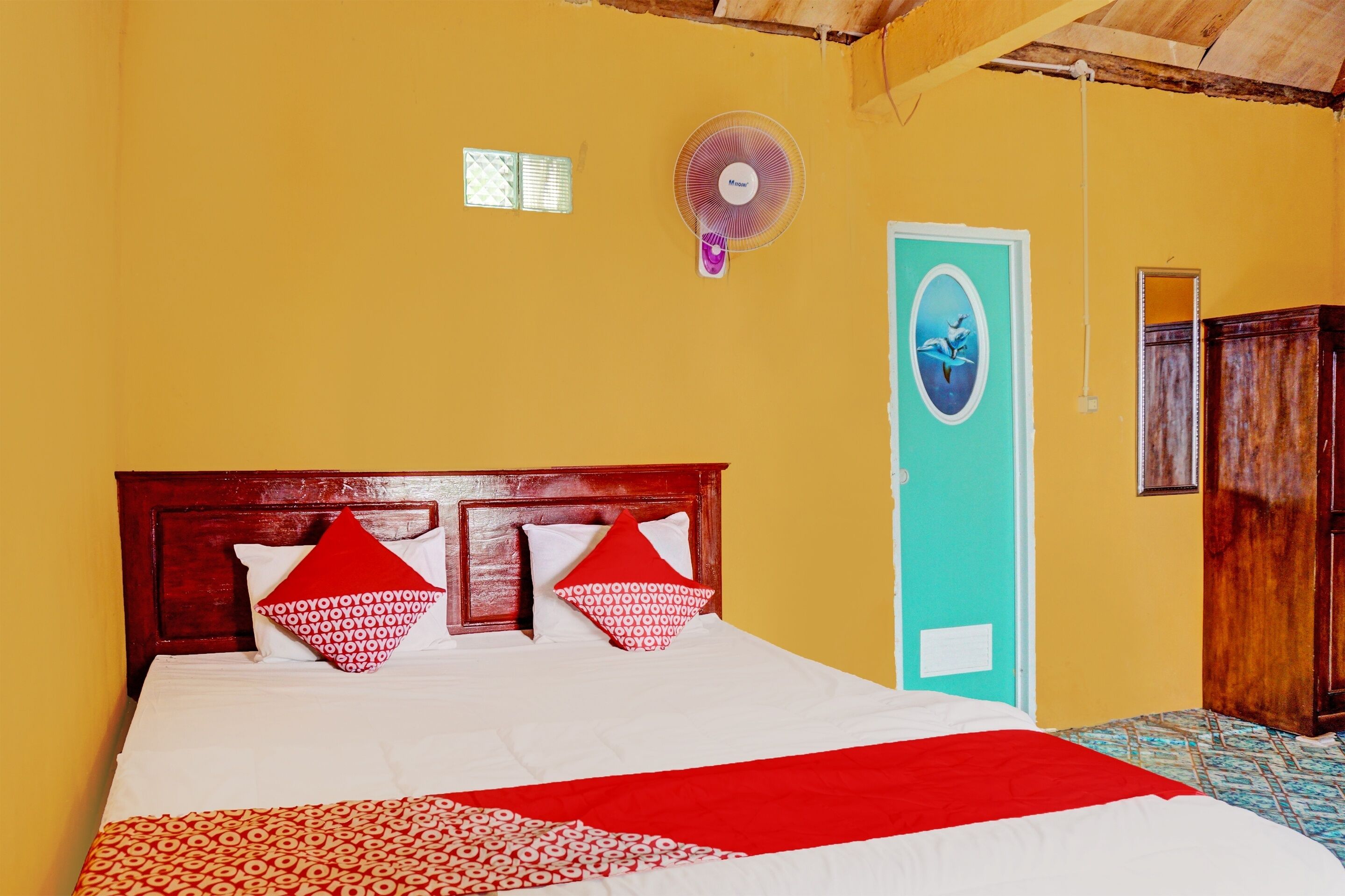 Un lit avec une tête de lit en bois, des oreillers à motifs rouges et blancs, et une couverture rouge. On y voit également un ventilateur fixé au mur et une porte avec un cadre bleu.