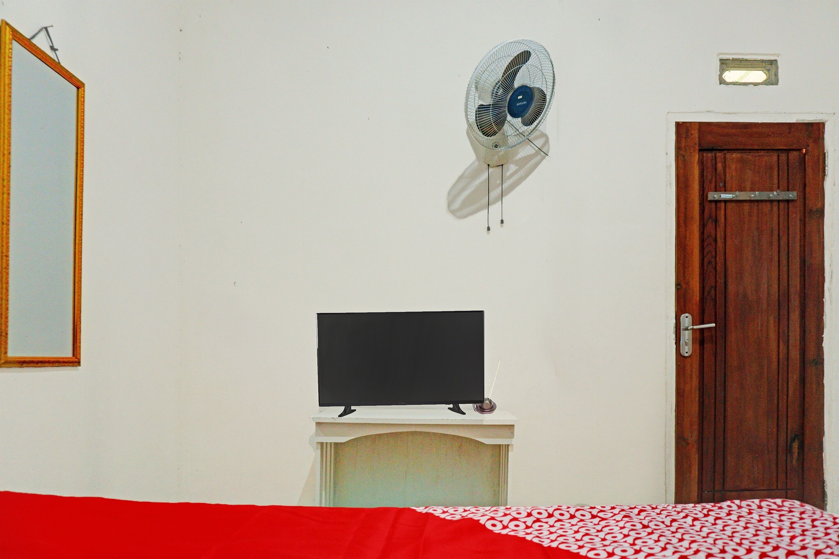 Ein Zimmer mit einem Bett, einem Fernseher auf einem Schrank, einem Wandventilator und einer Holztür.