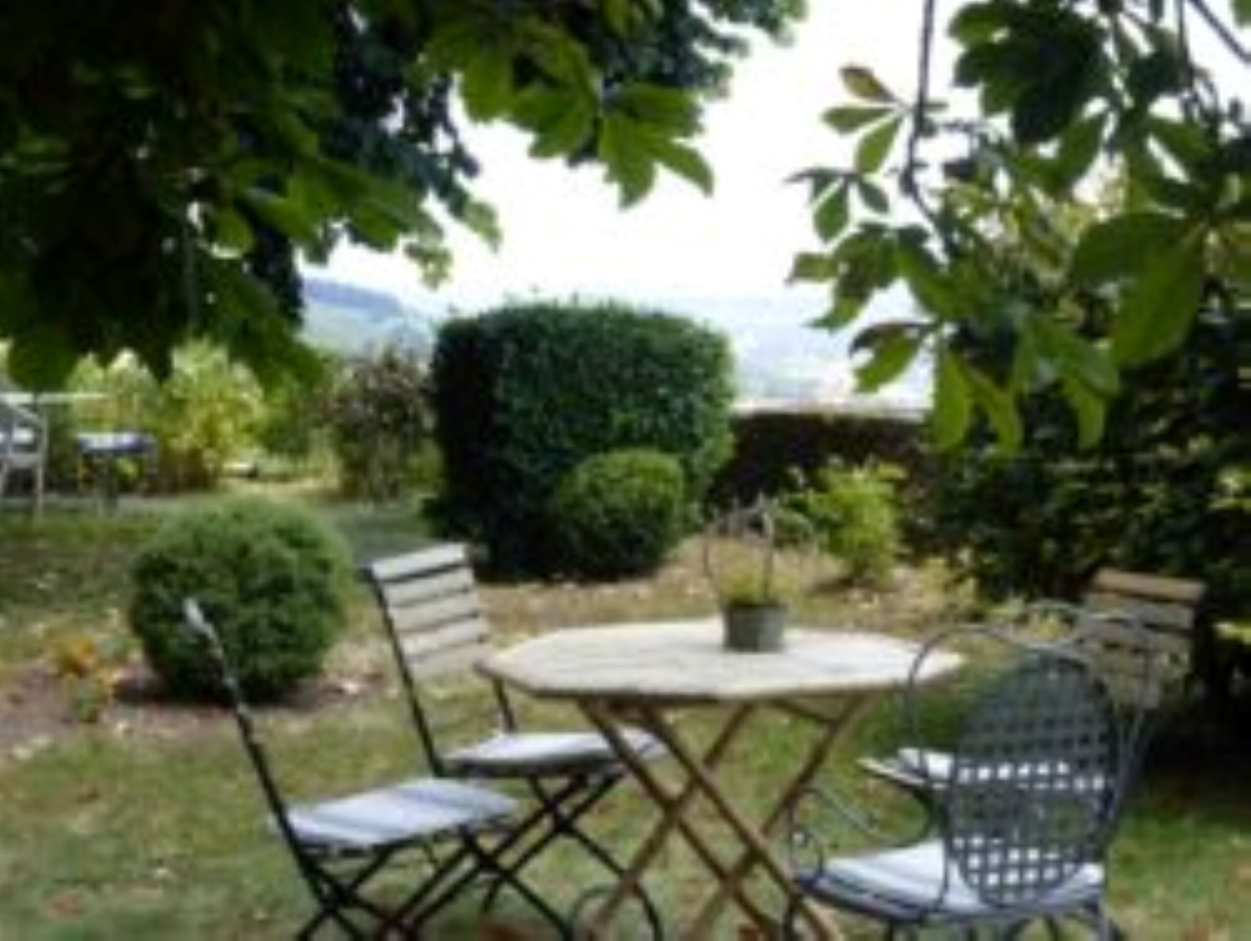 Un espace extérieur aménagé avec une table ronde et quatre chaises, entouré de verdure et d’une haie.