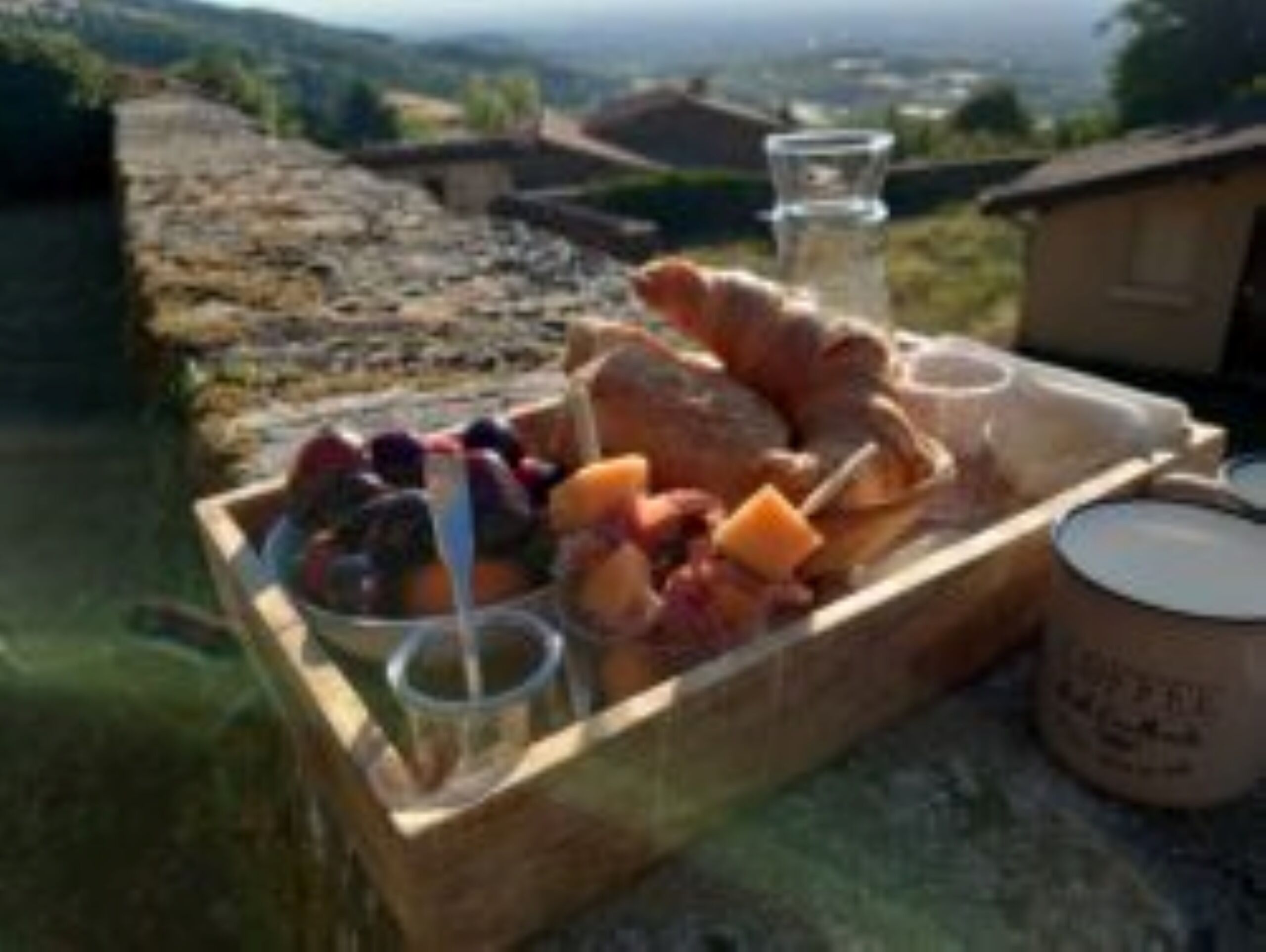 Uma caixa de madeira com diversos queijos, frutas e um copo de água numa varanda com uma vista panorâmica.