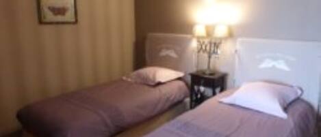 A room with two single beds, a nightstand with a lamp, and a wall-mounted shelf.