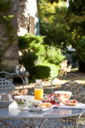 Mesa de comedor al aire libre con comida y bebidas, rodeada de follaje y edificios.