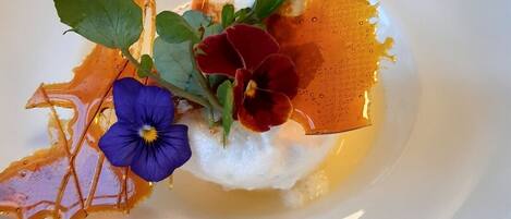 A white plate with a dessert featuring a dollop of cream, a purple flower, a red flower, green leaves, and a drizzle of orange syrup.