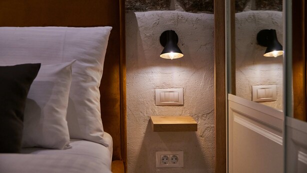 Un dormitorio con cama, mesita de noche, luz empotrada en la pared, toma de corriente y estante de madera.