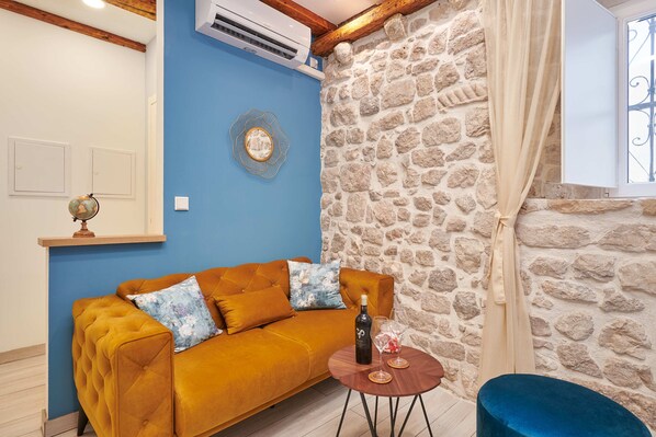 A living room with a blue wall, a stone wall, a wooden ceiling, a sofa, a small round table with a bottle and glasses, and a curtain.