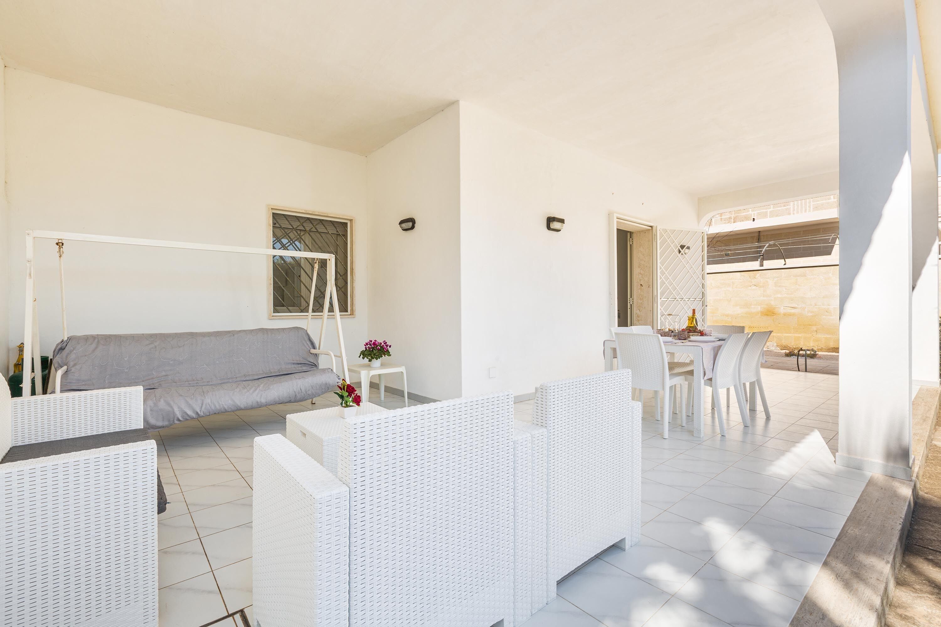 Una habitación moderna y bien iluminada con un sofá, muebles de mimbre blanco, un comedor con mesa y sillas, y vistas al exterior.