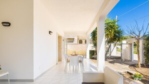 Un patio cubierto con mobiliario de mimbre blanco, una mesa de comedor y vista a un edificio al fondo.