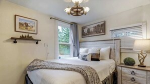 A bedroom with a large bed, a nightstand with a lamp, a window with blinds, and two framed pictures on the wall. - Come to the Hideout! Hot Tub, Fire Pits & Views! (Woodland Park)