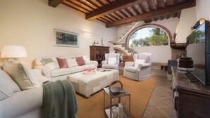 A spacious living area with a white sofa, two armchairs, a coffee table, and a fireplace. A staircase leads to an upper level, and a large window provides natural light.