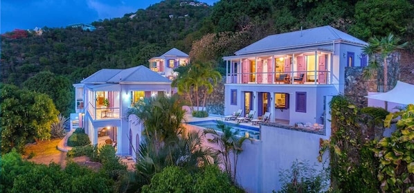 A multi-story house with a pool, surrounded by lush greenery and hills.