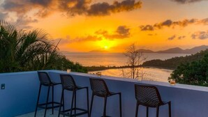 A balcony with three chairs, a view of the sunset over the sea, and a distant mountain range.