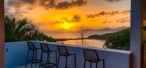 A balcony with three chairs, a view of the sunset over the sea, and a distant mountain range.