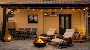 An outdoor seating area with chairs and a fire pit, a sign reading "Ferienwohnung Hofidyll," and a door with a green frame. - Vacation apartment "Hofidylle" on the pasture farm (Beelitz)