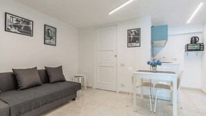 Un salon moderne avec un canapé gris, une petite table blanche et deux chaises. On peut voir des tableaux encadrés au mur, une porte blanche et un coin cuisine équipé d’un micro-ondes et d’une bouilloire.