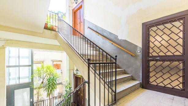 Un escalier avec une rampe en métal, une porte en bois ornée d’un motif en treillis et des plantes en pot sur le palier.