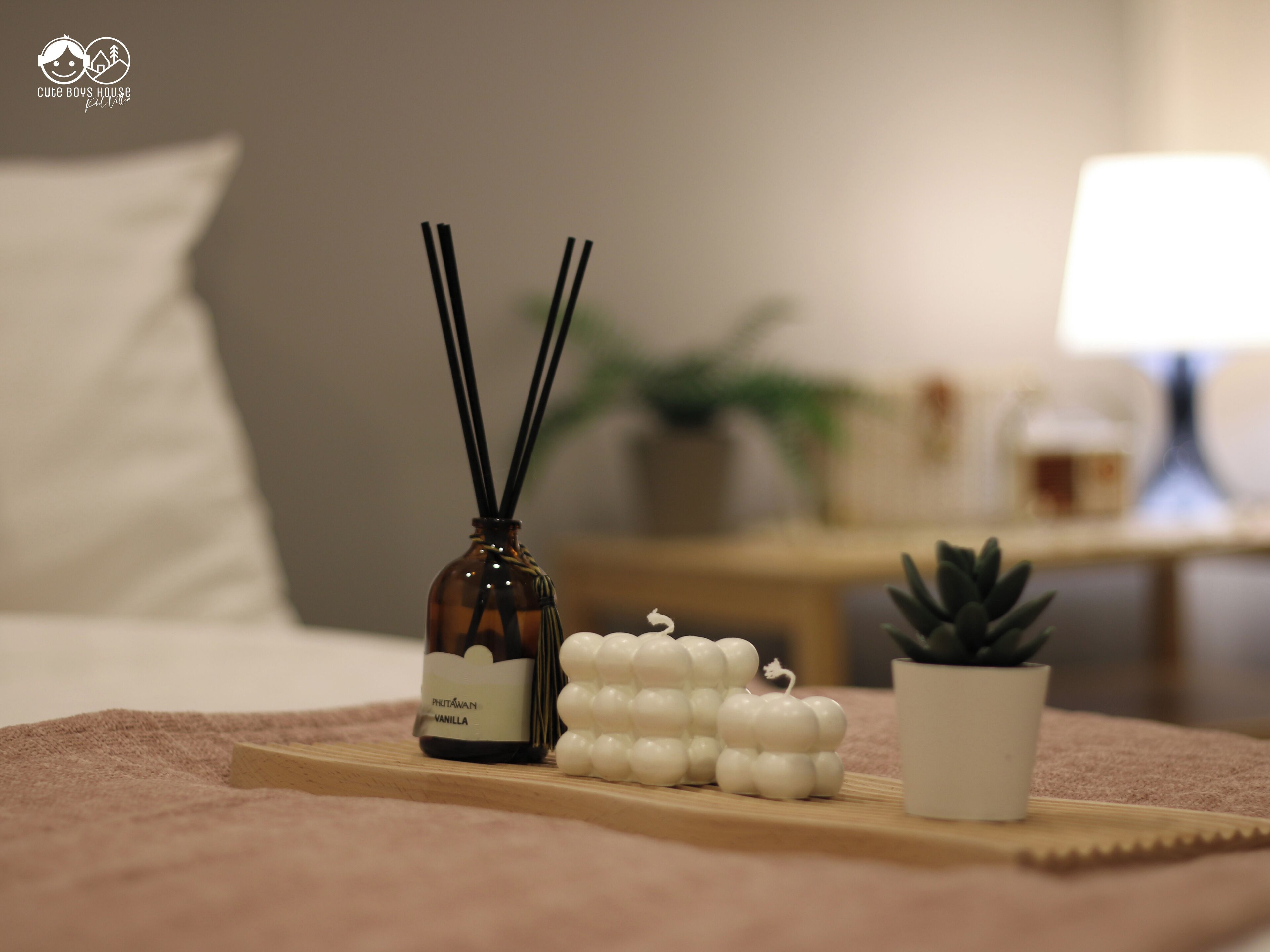 A bedroom with a bed, a bedside table with a lamp, a small potted plant, and a tray with a reed diffuser and candles.