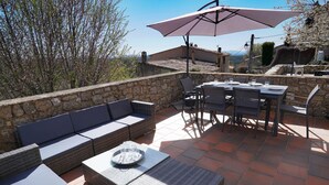 Een terras buiten met stenen muren, een rieten bank, een eettafel met stoelen en een grote parasol.