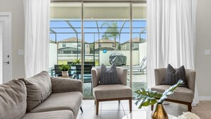 A living room with a sofa, two armchairs, a coffee table, and a view through a glass door to an outdoor patio area with a grill and chairs.