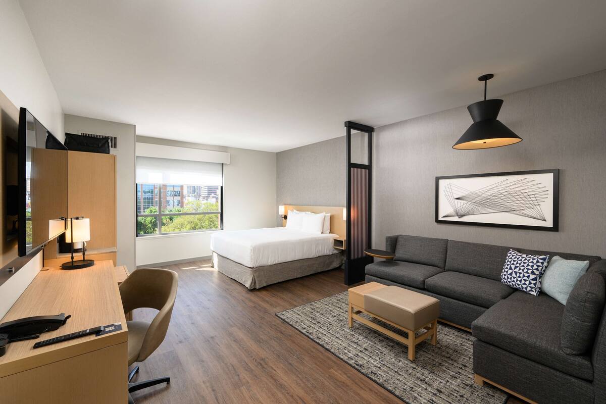A modern hotel room with a bed, sofa, desk, and a large window.