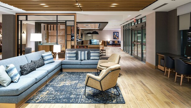 A modern hotel lobby with a sectional sofa, a small round table, and a lounge chair. There is a wooden ceiling with recessed lighting, a large window, and a glass partition wall. - Hyatt Place Albany/Downtown (Albany)