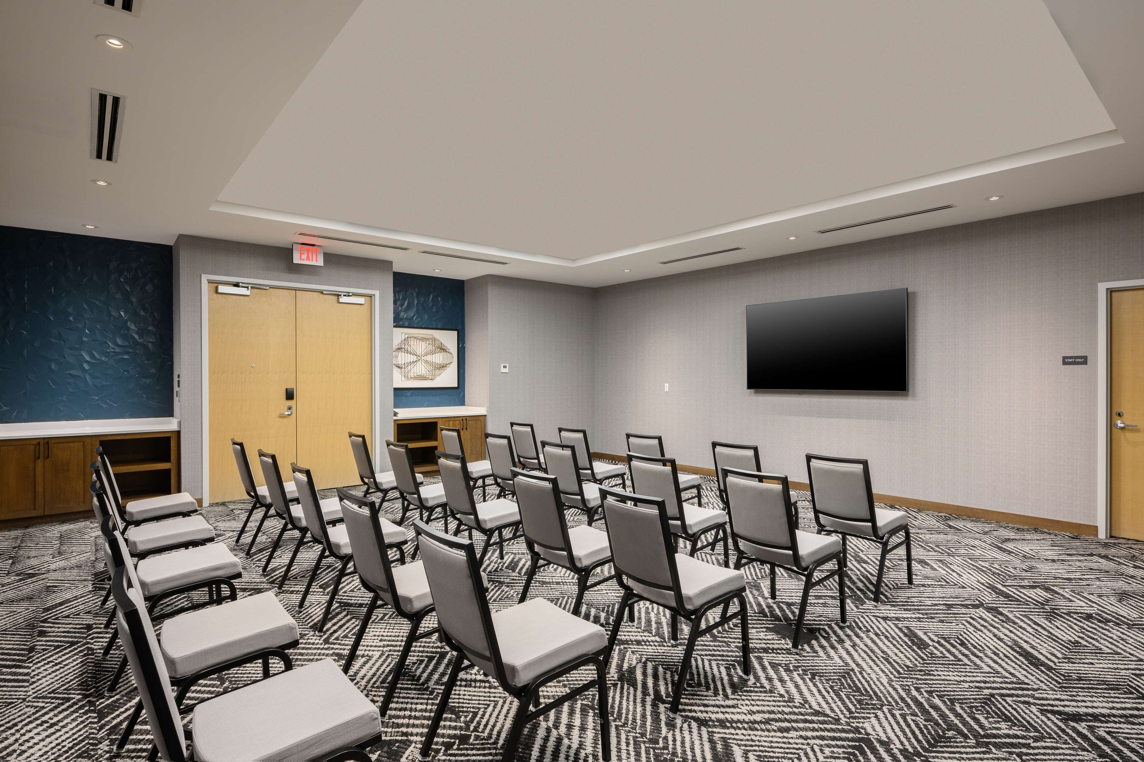 Uma sala de conferências com fileiras de cadeiras, uma tela grande e uma porta com sinal de saída.