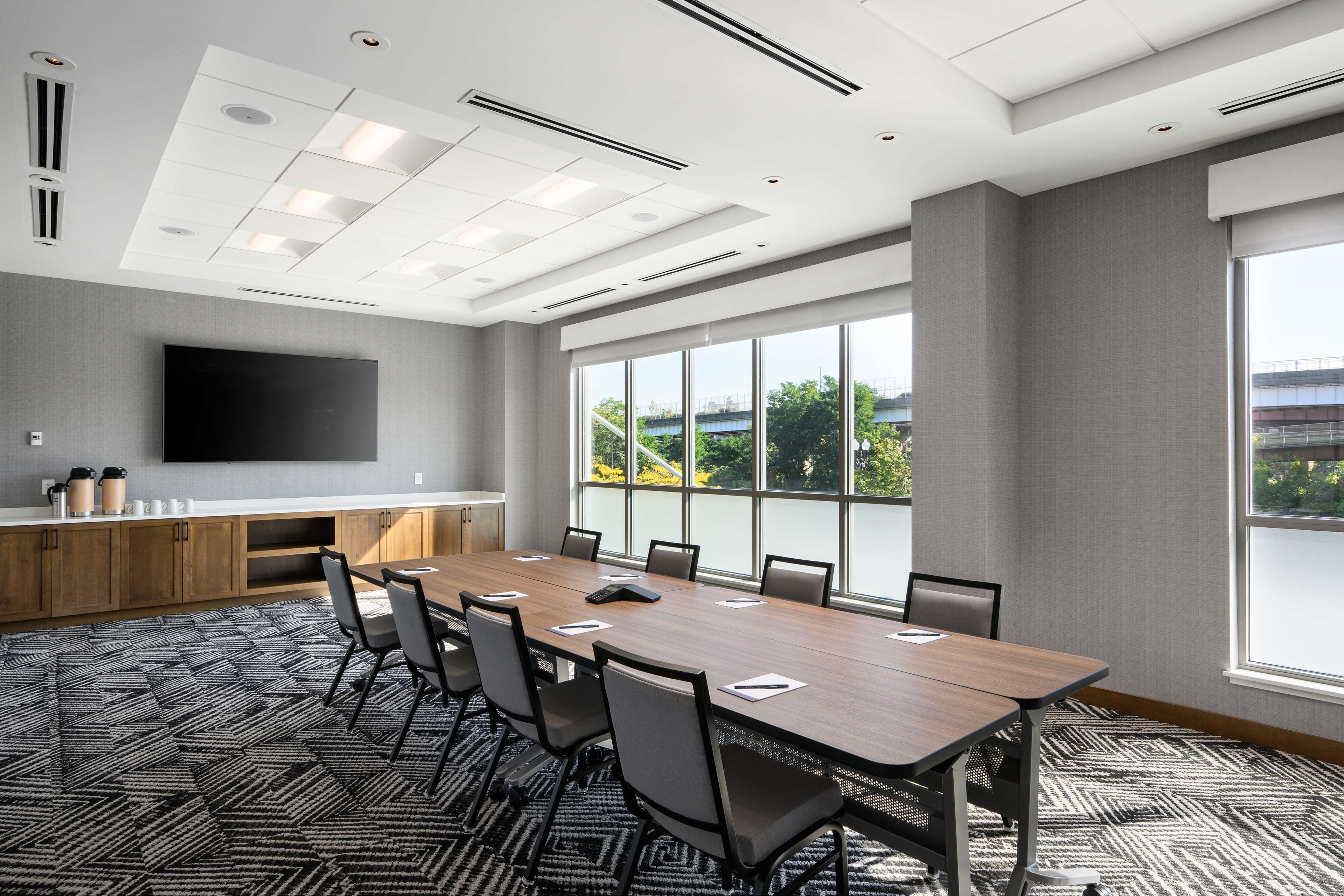 A conference room with a long table, chairs, a flat-screen TV, and large windows with a view of greenery.