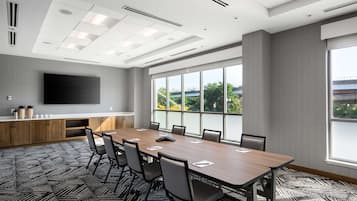 Un salón de conferencias con una larga mesa, sillas, un televisor de pantalla plana y ventanas amplias que dan a zonas verdes.