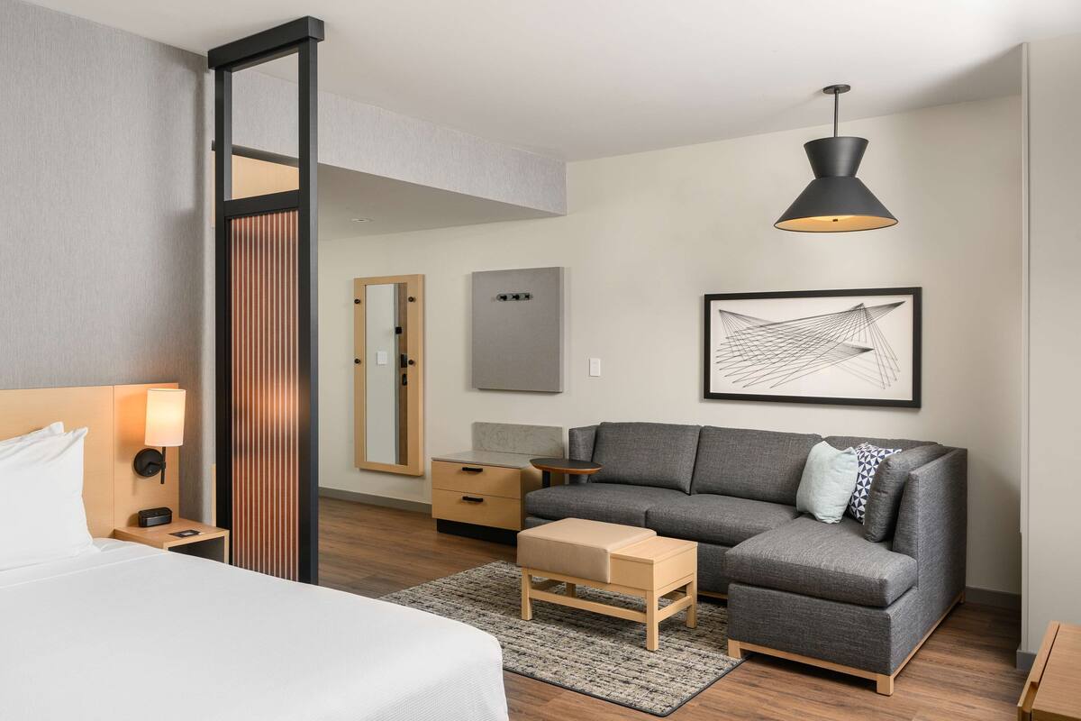 A modern hotel room with a bed, a grey sofa, a wooden nightstand, a small table, a floor lamp, a mirror, and a framed artwork on the wall.