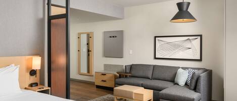 A modern hotel room with a bed, a grey sofa, a wooden nightstand, a small table, a floor lamp, a mirror, and a framed artwork on the wall.