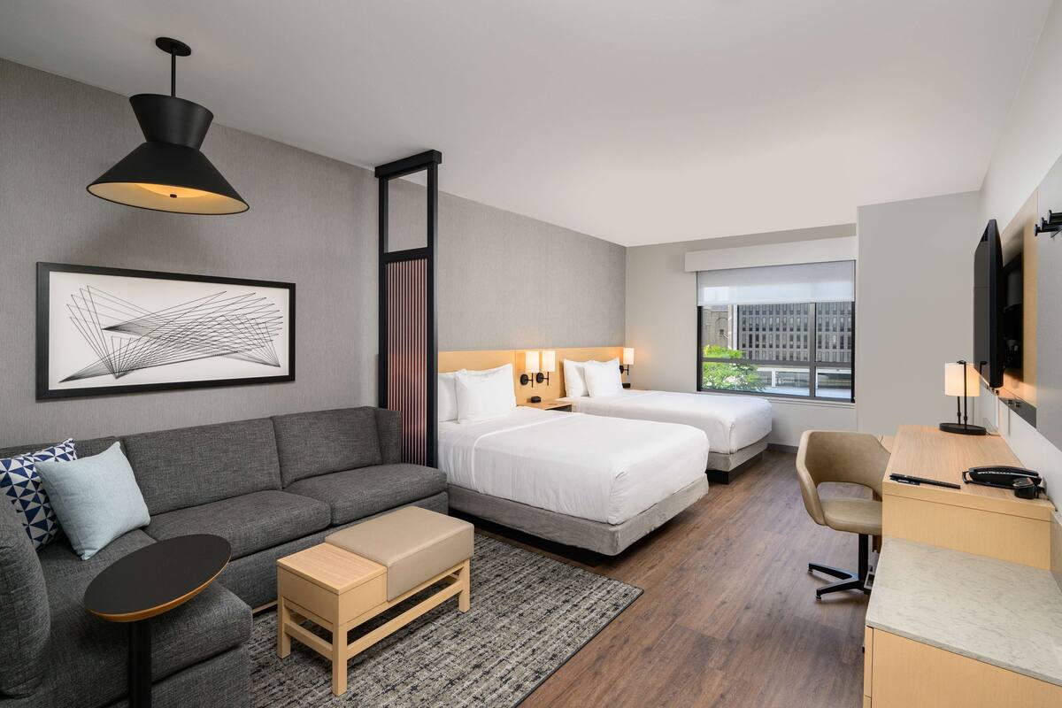 A modern hotel room with a grey sofa, two single beds, a desk, and a large window.