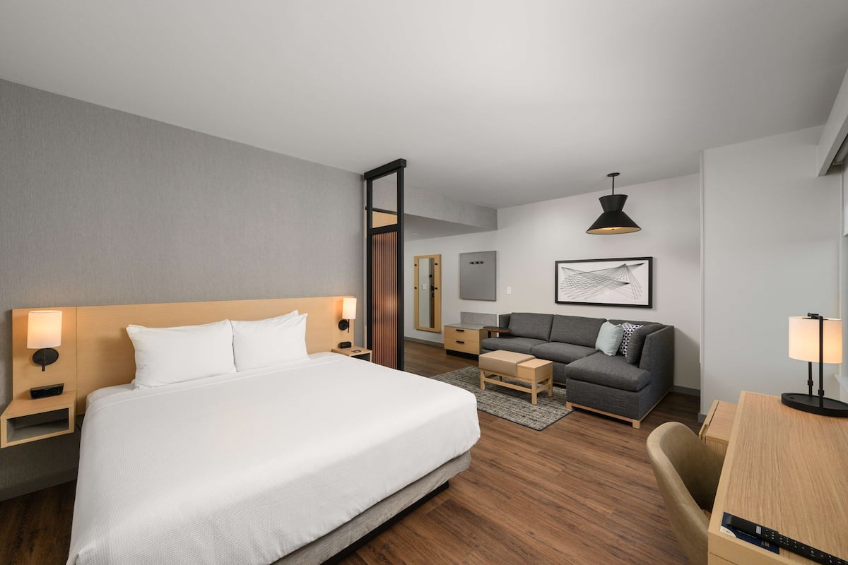 A modern hotel room with a large bed, a sofa, a desk, and a chair. There is a nightstand with a lamp, a mirror, and a small table. The room has a wooden floor and a gray wall.
