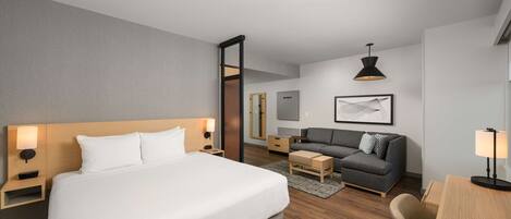 Una habitación de hotel moderna con una cama grande, un sofá, un escritorio y una silla. Se ve un mesita de noche con una lámpara, un espejo y una pequeña mesa. El piso es de madera y la pared es gris.