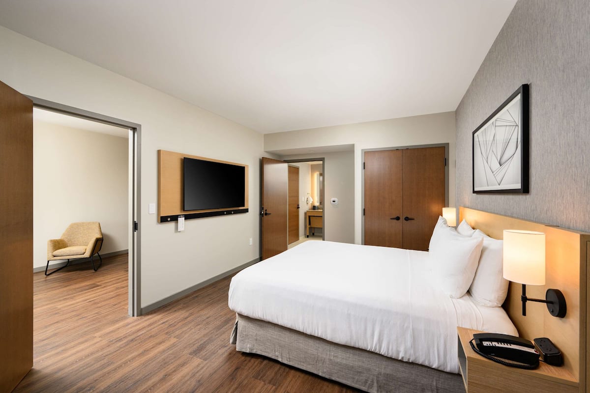 A hotel room with a large bed, a TV mounted on the wall, a desk with a lamp, and a chair.