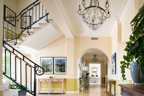 Ein imposantes Foyer mit Treppe, einer Chandelierleuchtre, gerahmten Kunstwerken und Blick in den Wohnbereich.