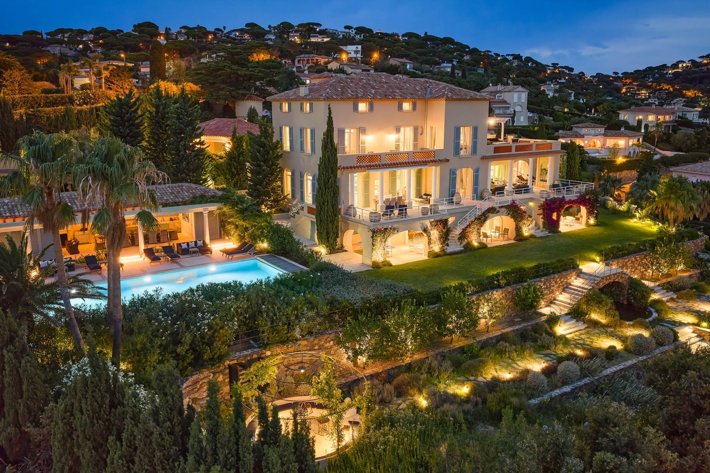 A large, multi-story house with a swimming pool, surrounded by lush greenery and a well-lit garden.