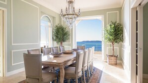 A dining room with a large table, chairs, and a chandelier. There are two potted plants and a view of the ocean through a large window.