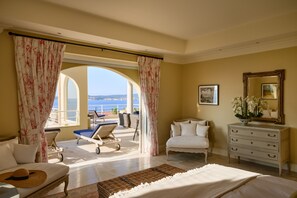 A hotel room with a bed, a sofa, a chaise lounge, a dresser, and a view of the sea through an arched doorway.