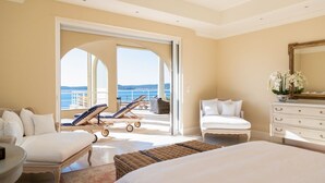 A spacious bedroom with a large bed, a sitting area with a chair and a small table, a white dresser, and a balcony with a view of the sea.