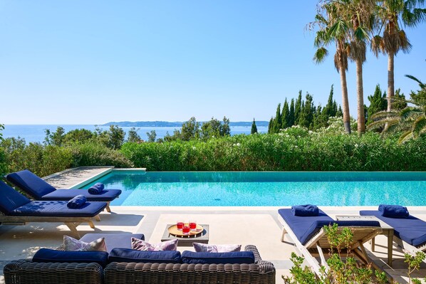 A poolside area with lounge chairs, a table with drinks, and a view of the sea and greenery.