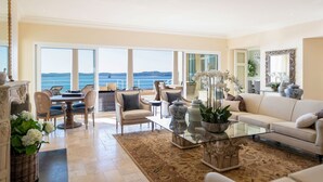 A spacious living room with a large window overlooking the sea, a comfortable sofa, a dining area, and a decorative fireplace.