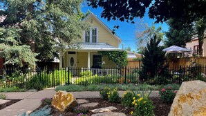 Una casa amarilla con porche, rodeada de un jardín con piedras para pisar, una cerca y diversas plantas.