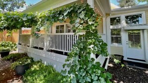Una casa con porche blanco, un espacio cubierto con enredaderas y un pequeño jardín con plantas en macetas.
