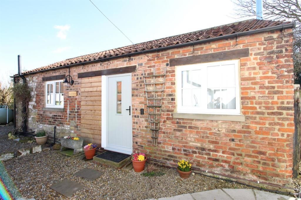 A small brick building with a white door, a window, and a ladder leaning against the wall. There are flower pots with flowers in front of the door.