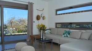 Ein modernes Wohnzimmer mit einer Couch, Poufs, einem Couchtisch und Blick auf Bäume durch große Fenster.