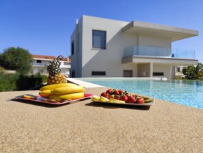 A modern poolside setting with a tropical fruit arrangement and a contemporary building in the background. - Home21-elegant Spacious Villa-5 min From the Beach (Rhodes)