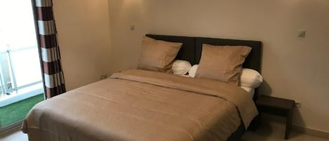 A bedroom with a large bed, two wall-mounted light fixtures, a small bedside table, and a view of a balcony with a striped curtain.