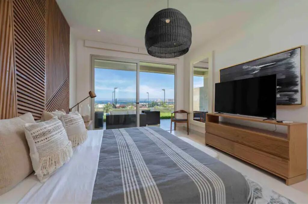 A modern bedroom with a large bed, a TV on a wooden stand, and a balcony with a view.