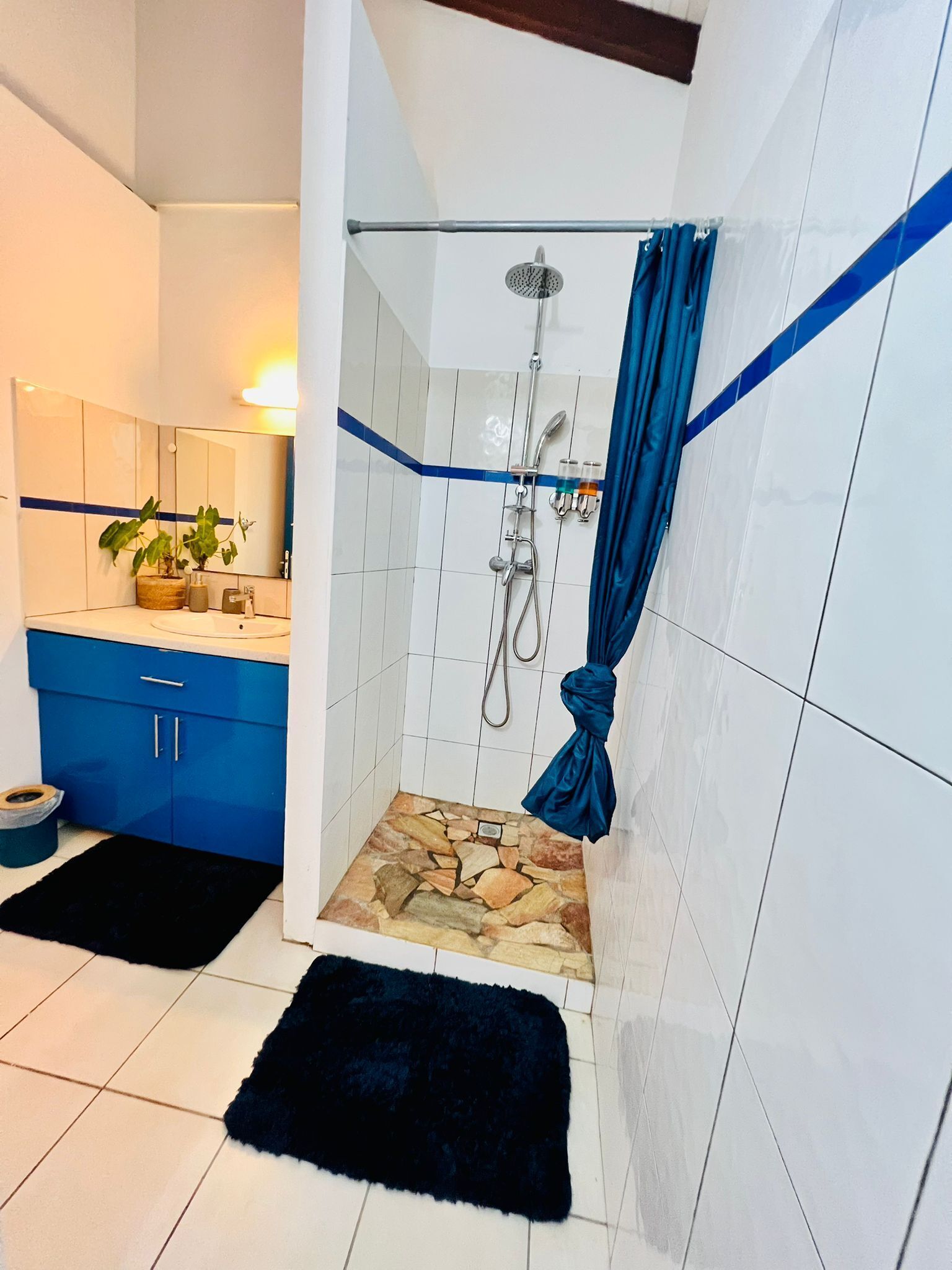 Une salle de bain avec une douche, un lavabo et un miroir. Il y a un rideau bleu et un tapis noir sur le sol.