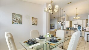 Een eetgedeelte met een glazen tafel, witte stoelen en zwart-wit geborduurde kommen. Er hangen schilderijen aan de muur en in de achtergrond is een keuken te zien.